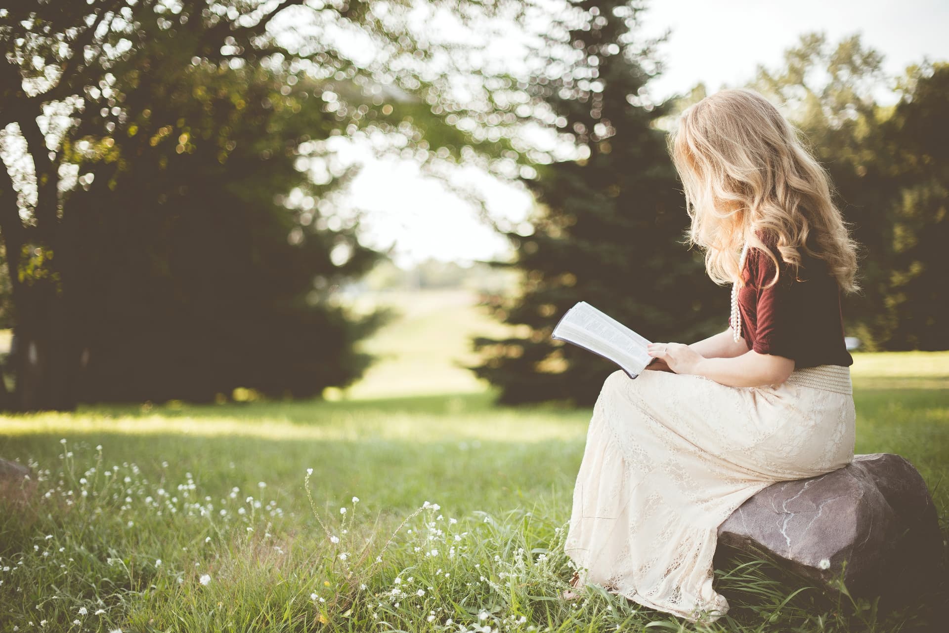 A woman reading her Bible on a rock in a sunlit meadow